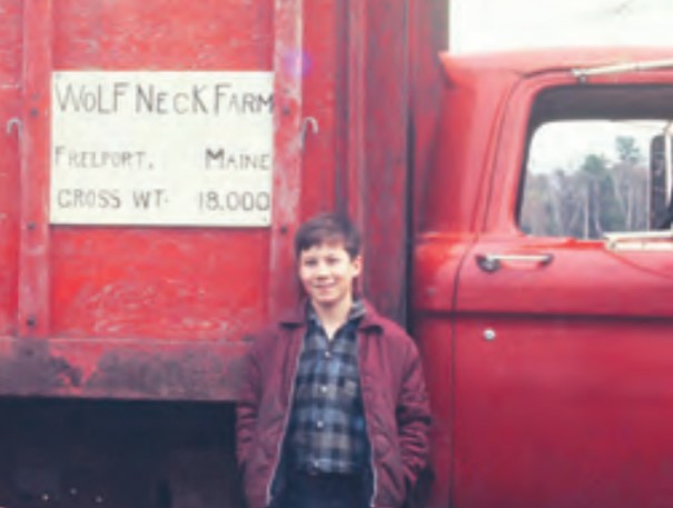 A boy standing in front of a red truck bearing a sign which reads "Wolf Neck Farm Freeport Maine Cross Wt 18,000