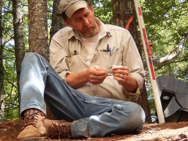 John Mann seated with legs crossed on a ledge in a forest with trees in the background. He is wearing a baseball cap, tan work shirt, and jeans. He is looking at a tool in his hands and beside him is a red and white striped pole used in surveying.