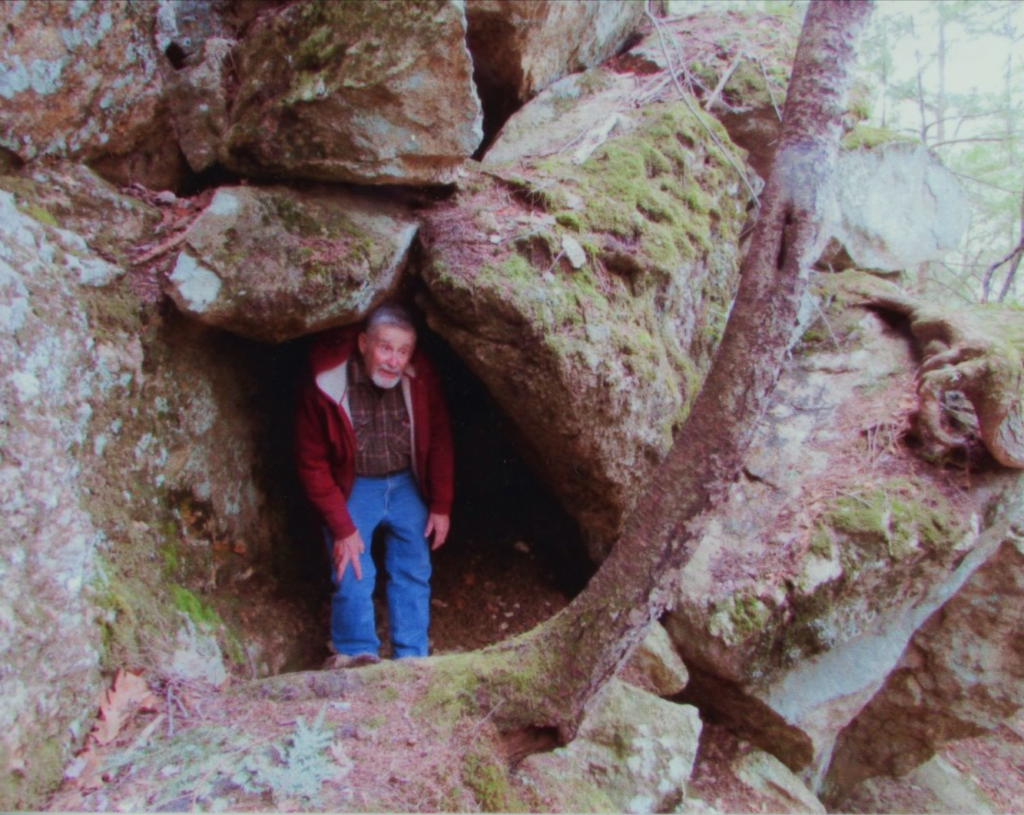 David Coffin stands in the entrance to a cave formed of large, moss covered boulders. He is wearing jeans and a red jacket.